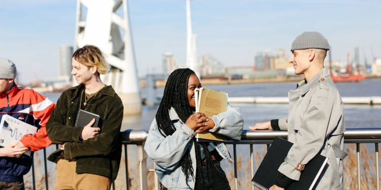 4 young students standing with their backs to the river thames, smiling and holding sketch books. The student in the centre of the image is holding a sketchbook up to her face and laughing.