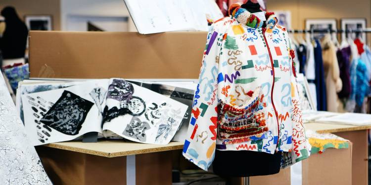A white jacket covered in brightly coloured patterns and lines on a mannequin. Sketchbooks and clothing rails on tables in the background