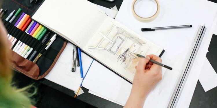 A hand holding a sketching pencil, drawing an living room in a small sketch book. There are brightly coloured pens and pencils to the left hand side of the sketchbook and the person illustrating only has their hand shown.