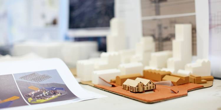 A small light brown wooden model of a house sits to the left next to computerised drawings printed