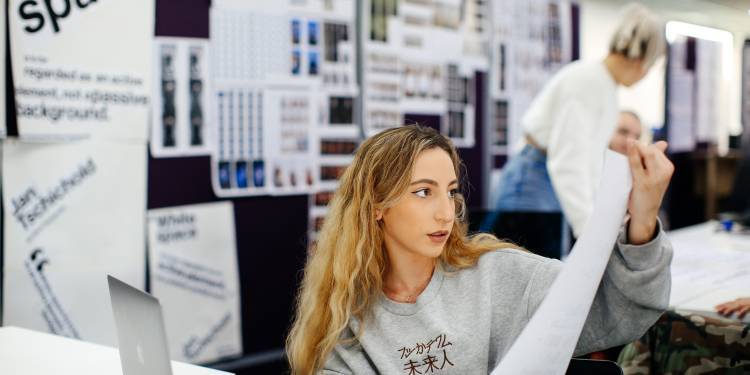 white woman with blonde hair holds large white paper with a laptop open