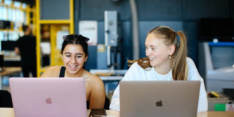 Two young woman work together on their laptops and smile
