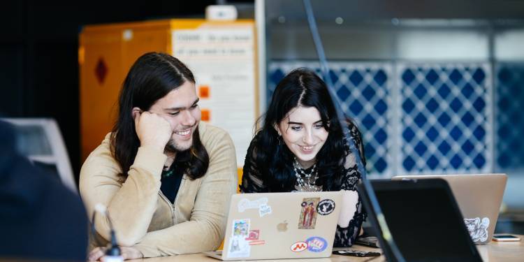 Two students smile while working together on their laptops.