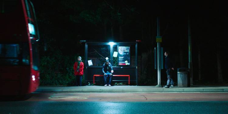 Film still of people standing at a bus stop in the dark with bus approaching to the left