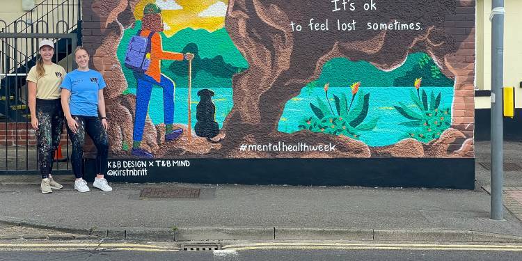 Kirsty and Brittany stand next to a mural they painted on the side of a building. The painting shows someone walking out of a cave to find a beautiful lake