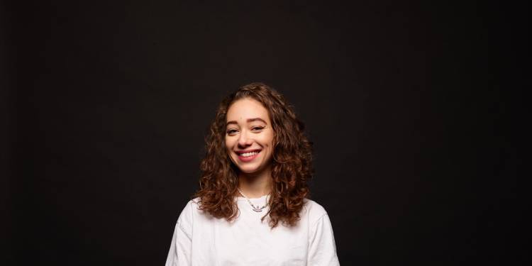 Young woman, early 20s, with curly brown hair smiles at the camera