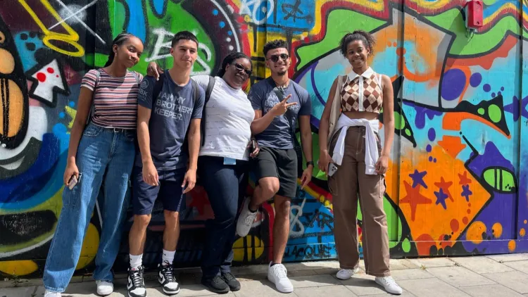 Five people lean against a graffited wall posing