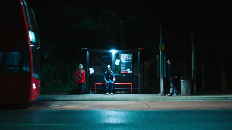 People stood at a dimly-lit bus stop with bus approaching to the left
