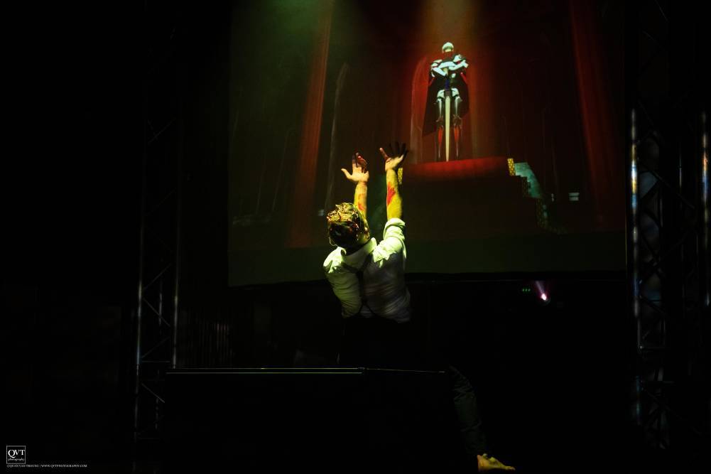 A performer in the dark castle stretches his hands up in front of a suit of armour