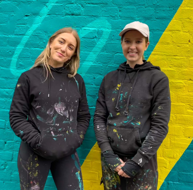 Two women stand in front of colourful mural wall