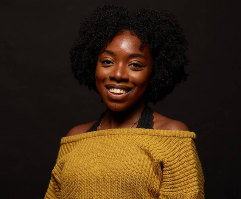 Professional-style photograph of female wearing a camel coloured off the shoulder jumper smiles for the camera against a black background