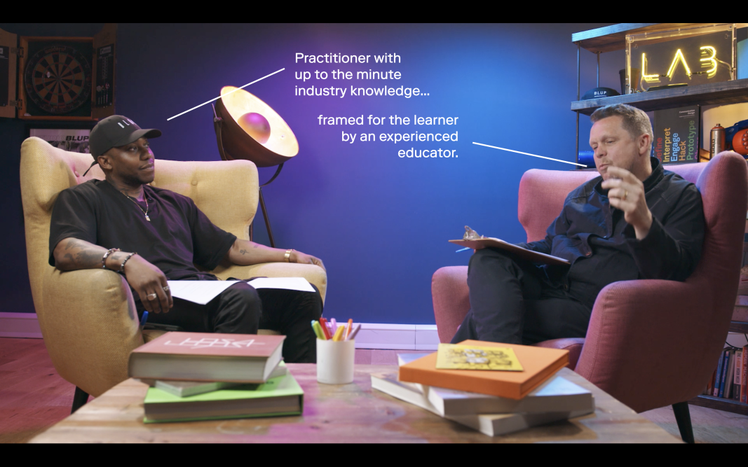 A black man wearing a black cap and tshirt sits in a large yellow armchair talking to a white man wearing a navy jacket sat on a red armchair, holding a clipboard