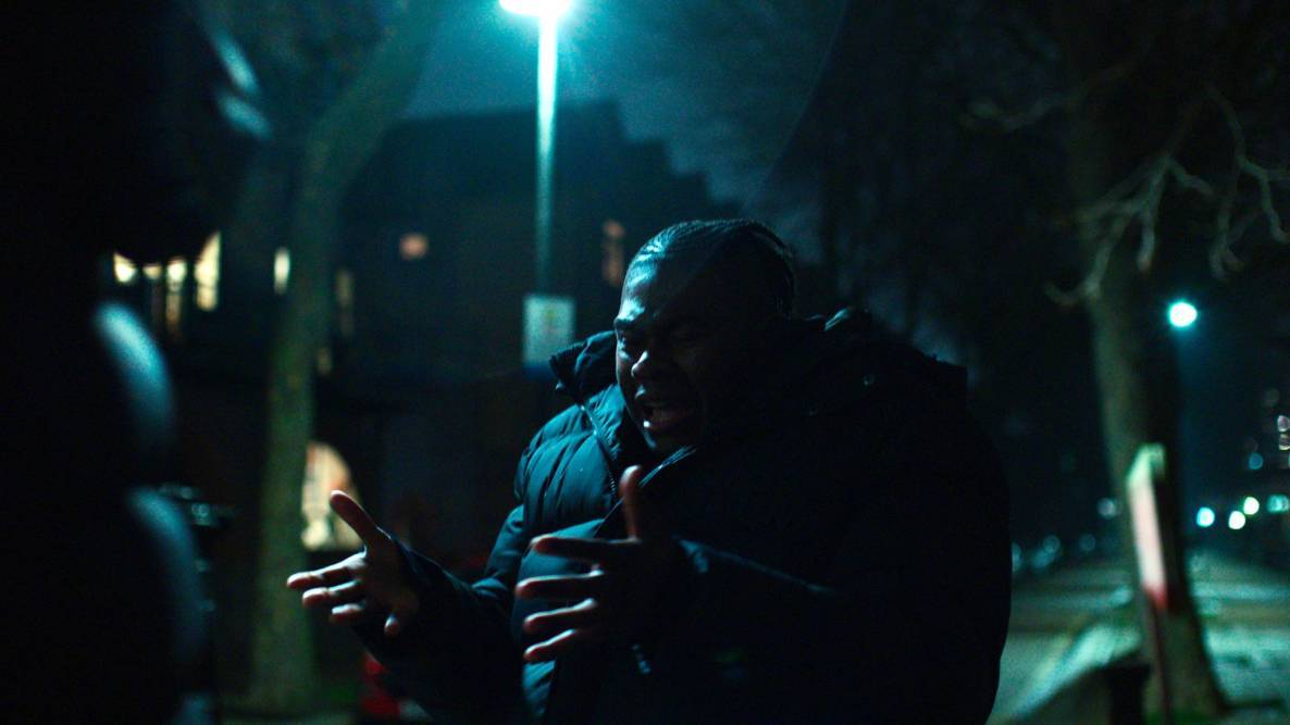 Man stood under lamppost in the night gestures with hands outstretched 