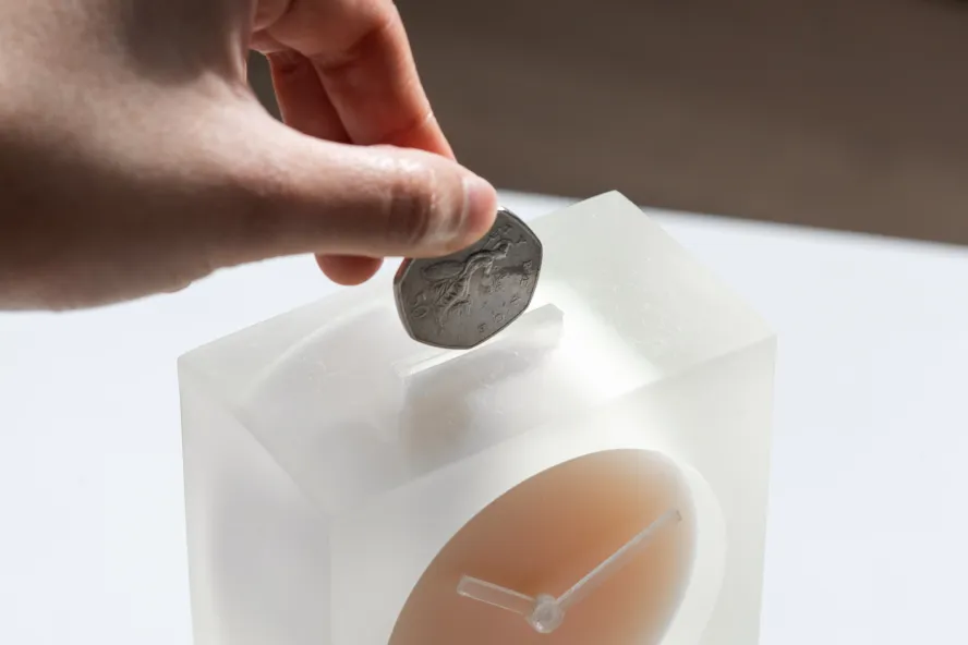 A hand putting 50 pence piece into a slot on the top of an analogue clock.