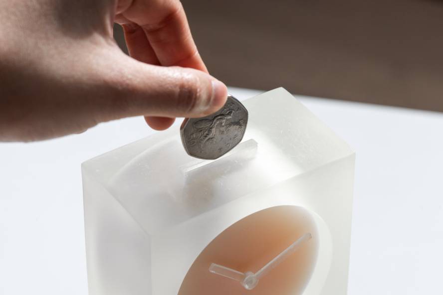 A hand putting 50 pence piece into a slot on the top of an analogue clock.