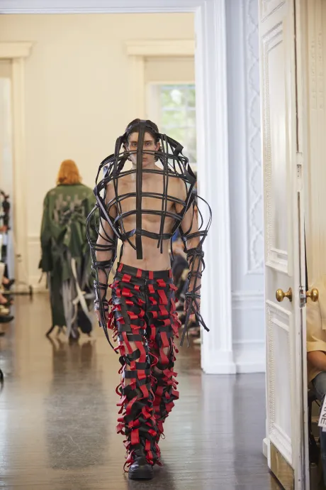 Male model wear a cage around his head and black trousers with red tags all over