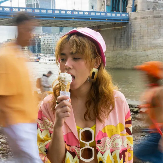 Girl wearing bucket hat and colourful shirt easting ice cream on the bank of the river Thames in London