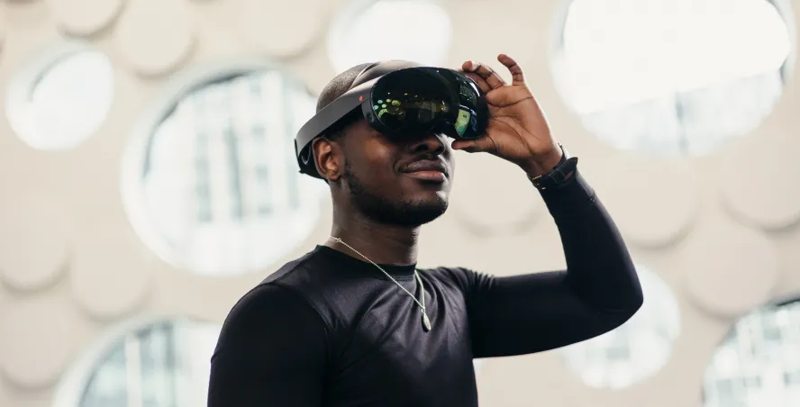 Student adjusts a virtual reality headset with his hand in a bright open space with circular windows