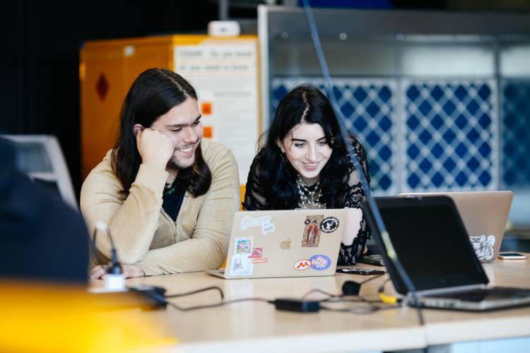 Two students smile while working together on their laptops.