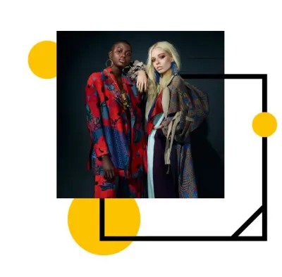 Two models in high fashion garments stand leaning on one another.