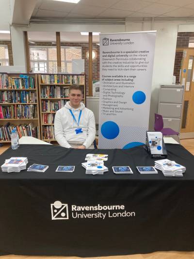 White man with blonde hair wears a white hooded jumper and a blue lanyard around his neck, sat a table with Ravensbourne branded print on