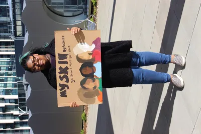 Young black woman standing with a painted placard with illustrated women of varying skin tones, with the slogan saying "my skin - not your business"