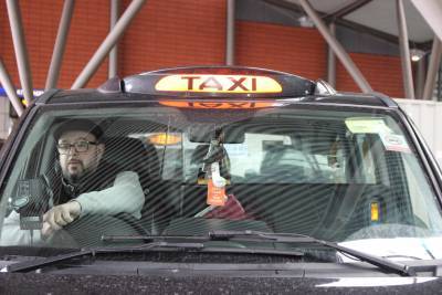 White taxi driver wearing square glasses and flat cap driving a black taxi, with its orange light on