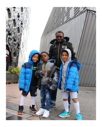 three young black children in bright blue coats stand posing with their dad behind smiling