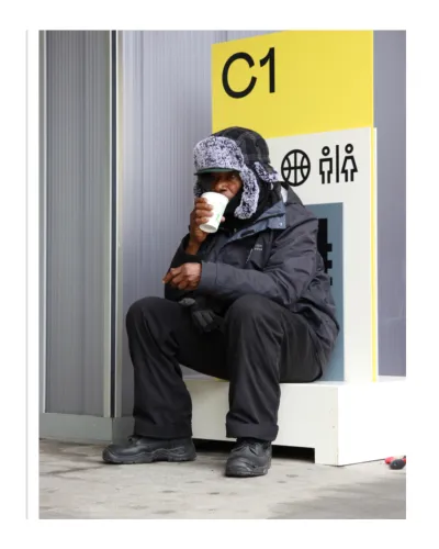 An older black man perches on a ledge drinking a coffee in a takeaway cup in front of a bright yellow sign.