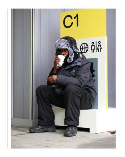 An older black man perches on a ledge drinking a coffee in a takeaway cup in front of a bright yellow sign.
