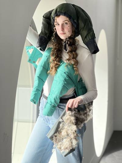 Young white women stands in front of a window wearing coats as different garments of clothing on her body.