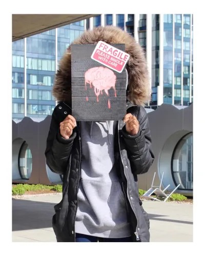 a faceless person stands holding a black placard with a painted pink brain melting on it, with a large fragile sticker on top.