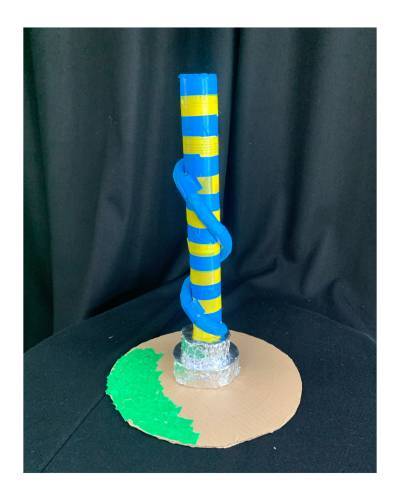 Small scultpture with blue and yellow striped cylinder stands tall with green cardboard base and a blue slide round the tall sculpture