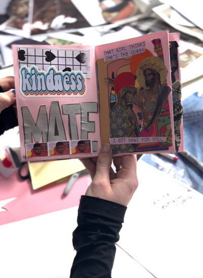 A white hand holds open a small pink booklet with stickers and collage all over it with "kindness mate" written in large bold writing on the left hand side