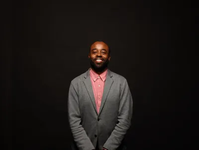 Man wearing a pink shirt and grey cardigan smiles to the camera