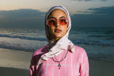 Woman wearing a head scarf, sunglasses and pink sweater looking into the camera with the sea shore in the background