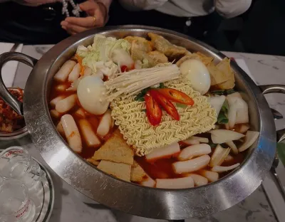 A large metal bowl contains noodles, rice cakes, chillis, eggs and vegetables on a table accompanied by a bottle of drink and glasses. 